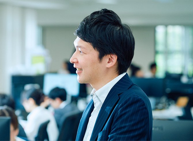 大人数のいるオフィスで笑顔で話を聞く男性社員